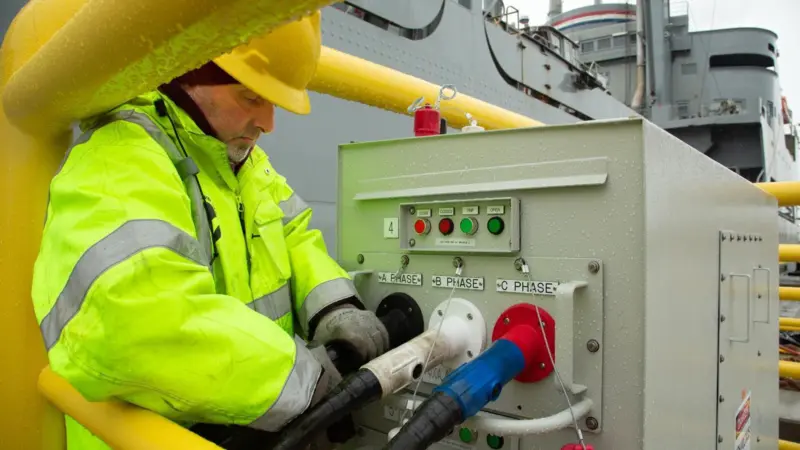 The image shows a man dressed in a yellow high vis connecting a vessel to shore power