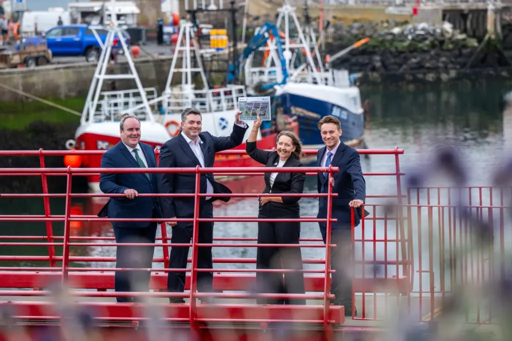 Four members of Eyemouth Harbour Trust standing on a red gangway holding aloft the harbour's masterplan