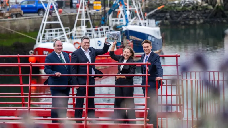 Four members of Eyemouth Harbour Trust standing on a red gangway holding aloft the harbour's masterplan