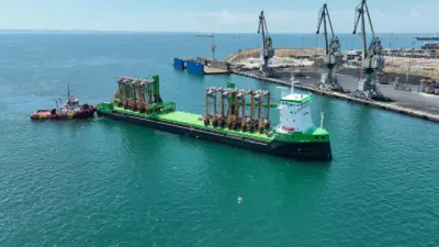 Eight hybrid straddle carriers arrive on a green barge at the Port of Thessaloniki
